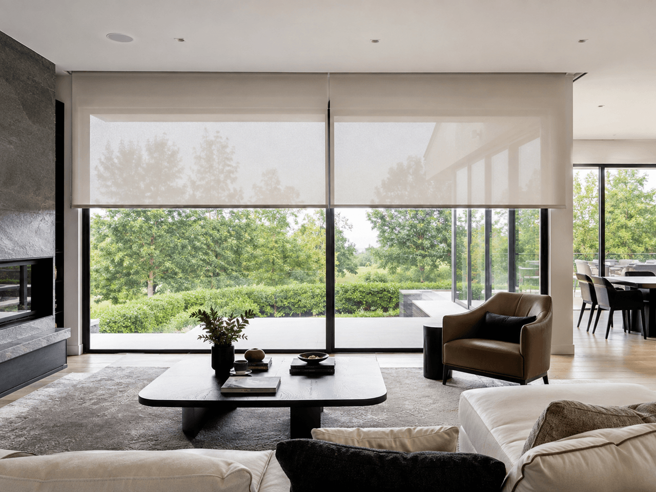 Contemporary living room with large glass doors, translucent motorized shades, a modern fireplace, minimalist furniture, and views of a lush garden