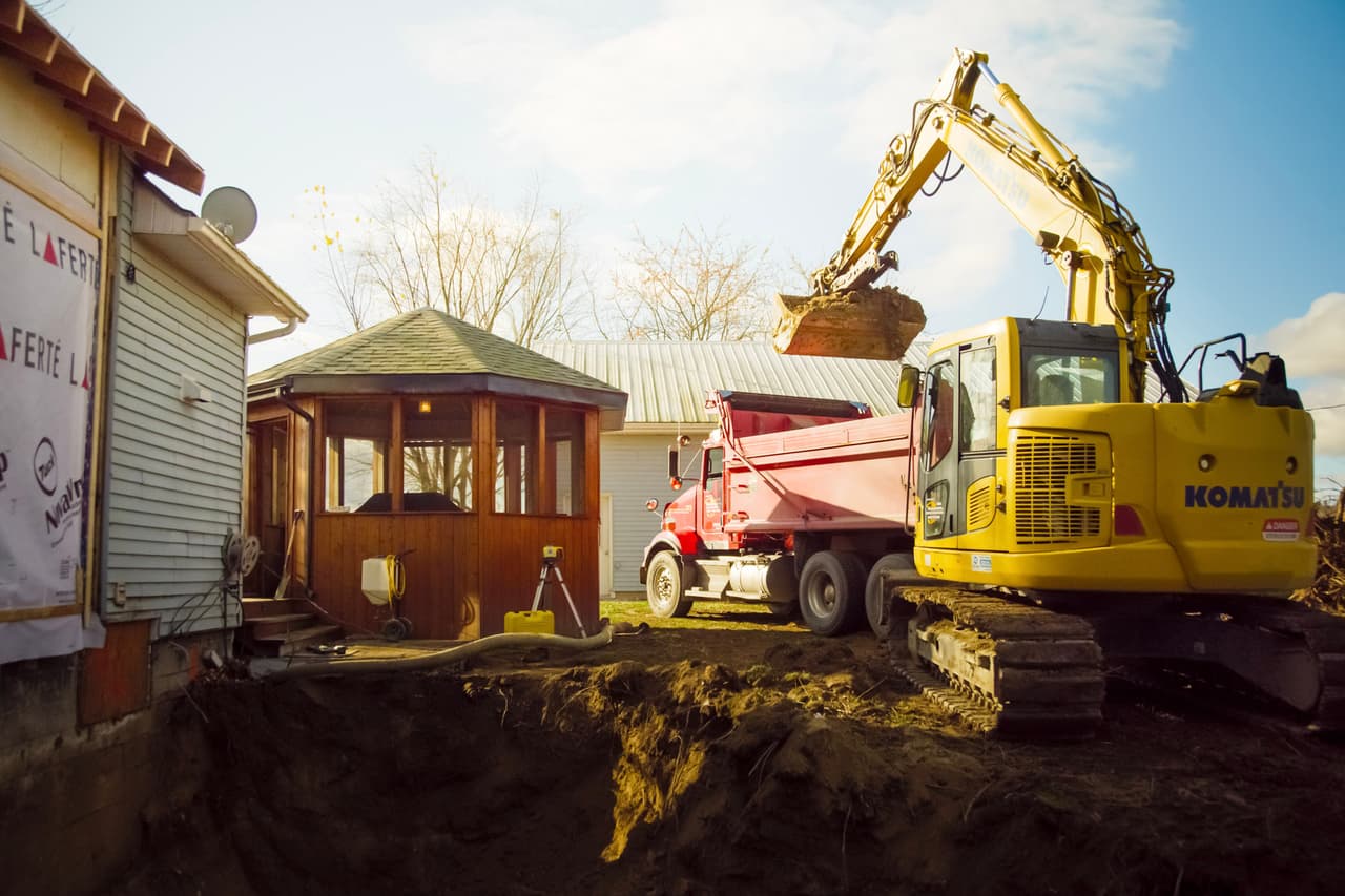 Komatsu excavator digging a pit near a house and a wooden gazebo, with a red dump truck ready to receive soil.
