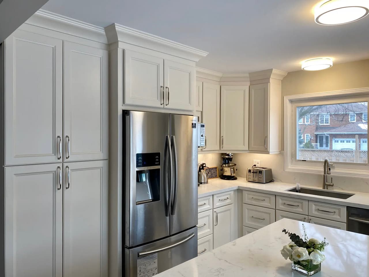 Modern and bright kitchen with full-height white cabinets, stainless steel refrigerator, marble-effect quartz counter, central island, under-window sink and ceiling lighting for a functional and elegant space