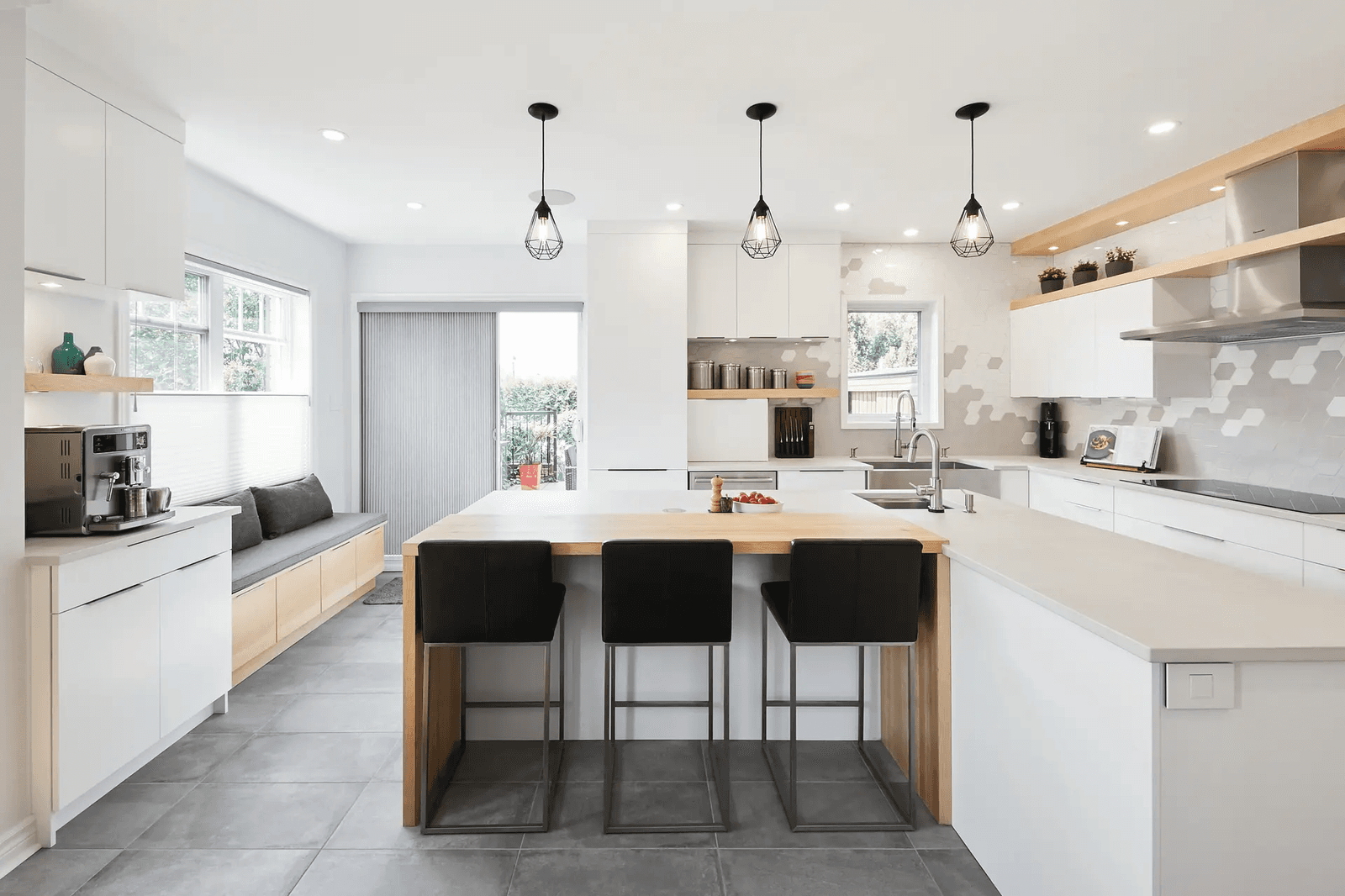 Scandinavian-style kitchen in Montreal featuring a spacious central island with black bar stools, minimalist white cabinets and a geometric mosaic backsplash. Grey ceramic flooring adds warmth to the space.