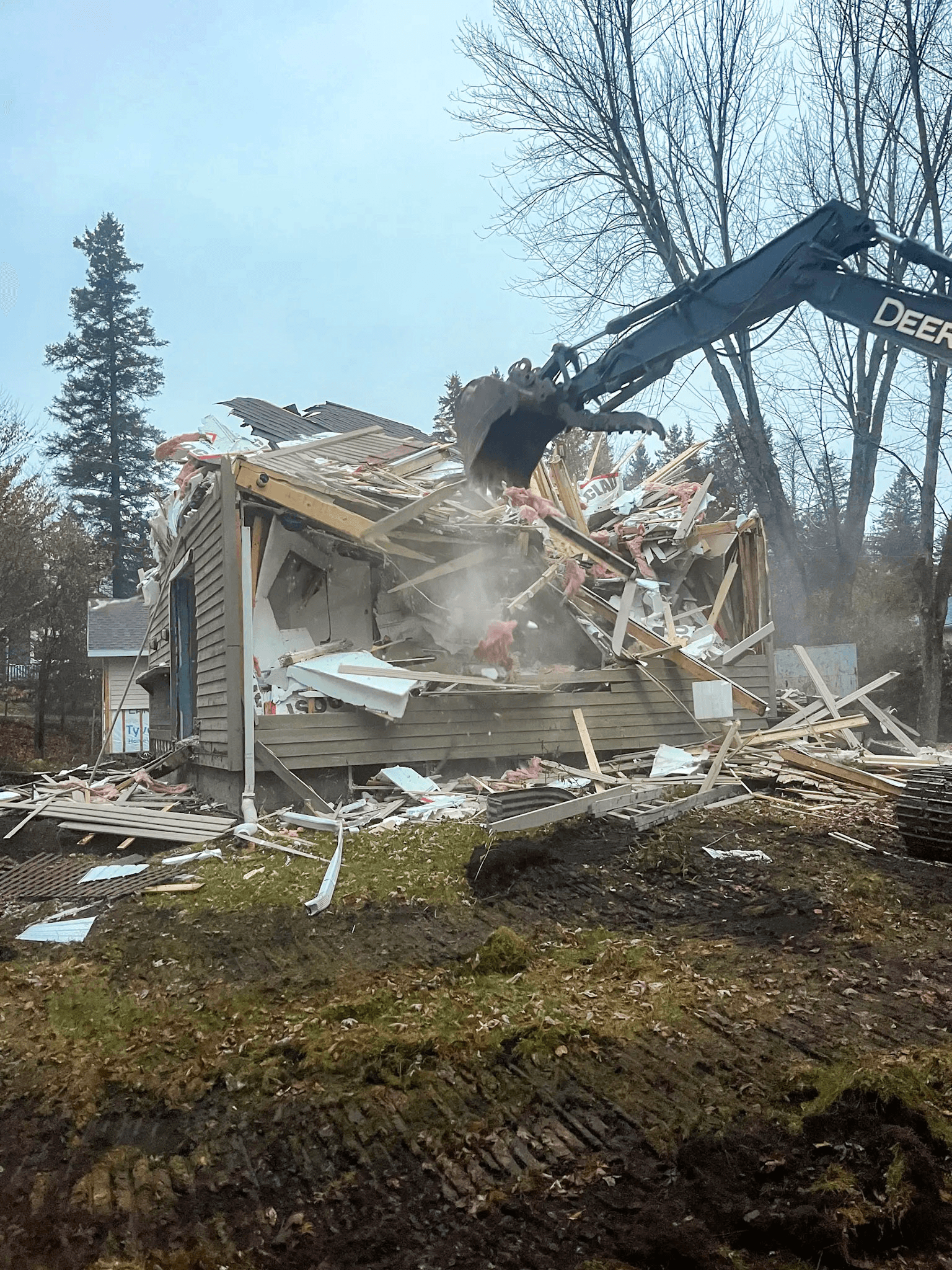 Demolition of a wooden house using an excavator in a residential area surrounded by trees.