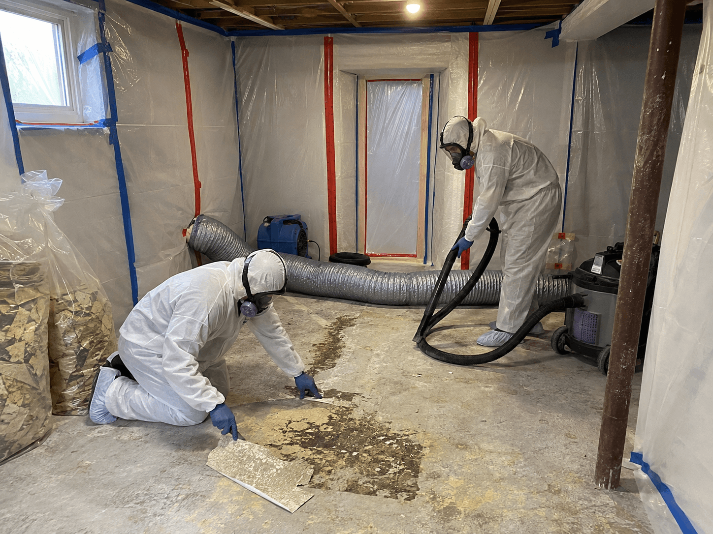 Workers in protective suits removing old flooring inside a contained work area with plastic sheeting, ventilation and an industrial vacuum