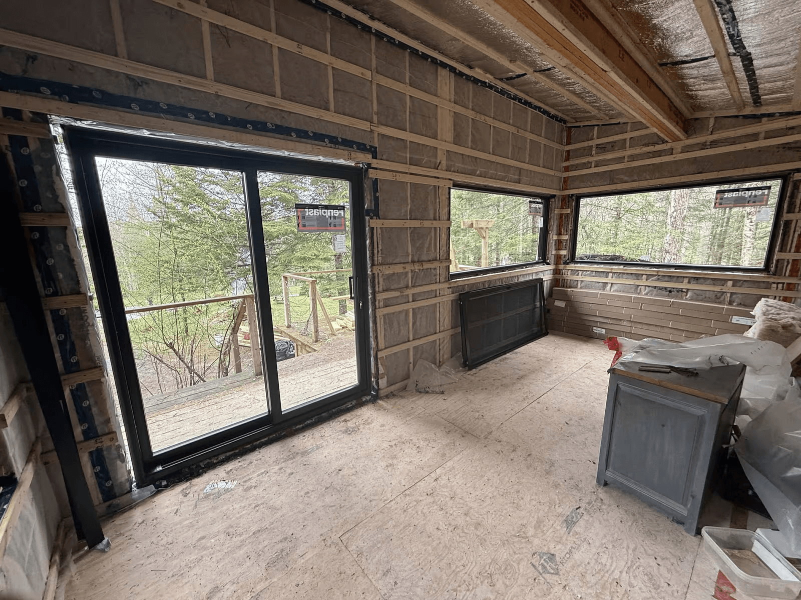 Residential room under renovation with large black patio door, panoramic windows, open walls with exposed insulation, wood framing and a view of a wooded property.