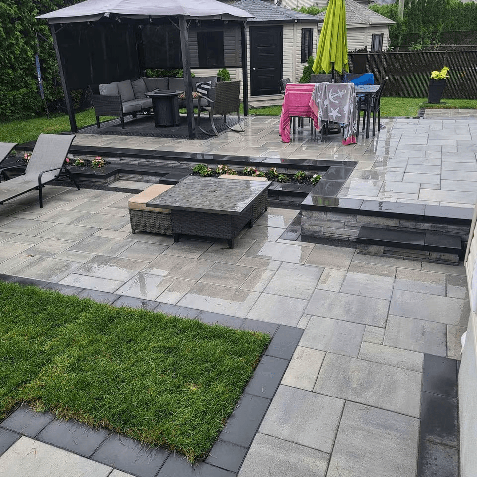 Landscaped backyard featuring a large grey interlocking stone patio, raised sections, stone steps, black edging, integrated planters, a screened gazebo, outdoor furniture, a green umbrella and a lawn.