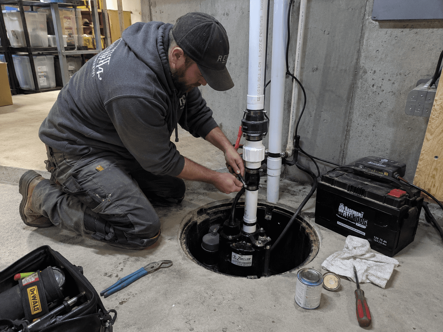 Technician installing and adjusting a sump pump in a basement with PVC piping, backup battery and plumbing tools