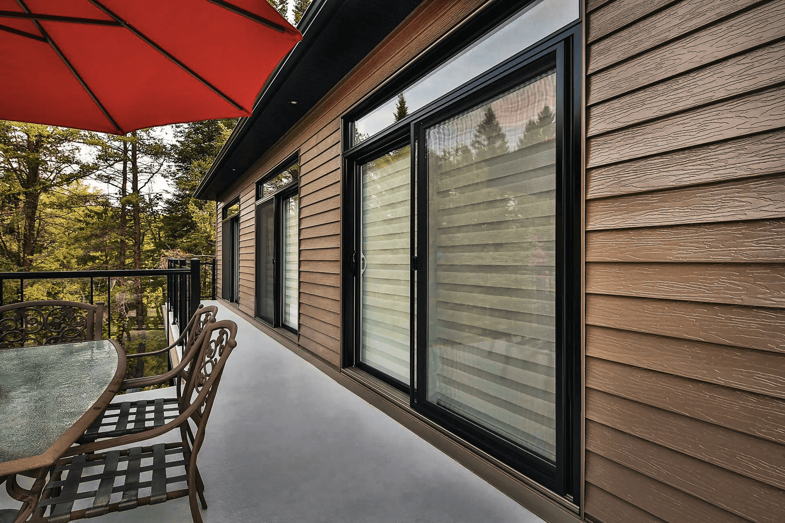 Exterior facade with large black windows, brown horizontal siding, furnished balcony with table and chairs, metal railing and view of trees.