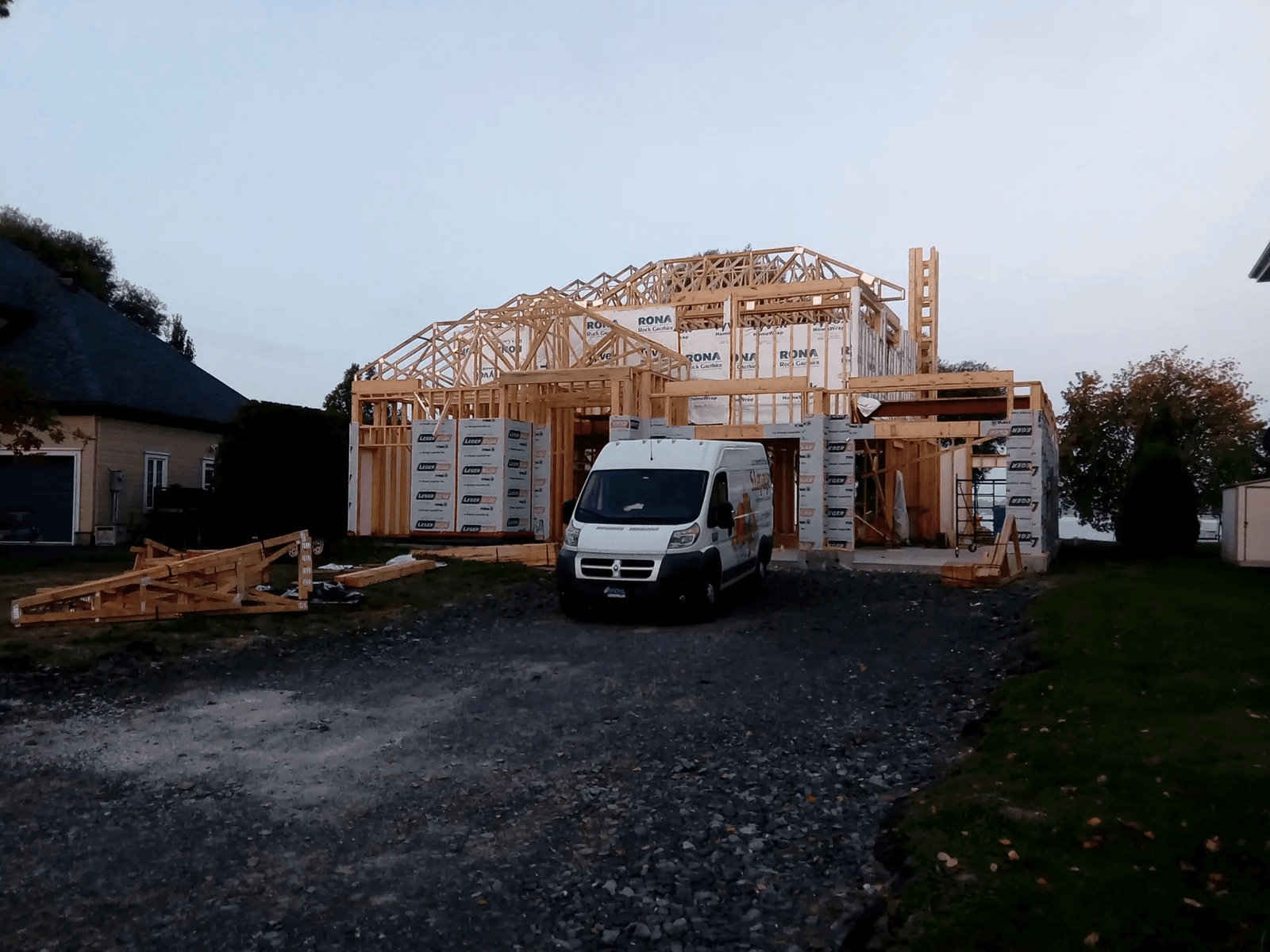 House under construction with wooden framing, RONA weather wrap, and a work van parked on the gravel driveway.