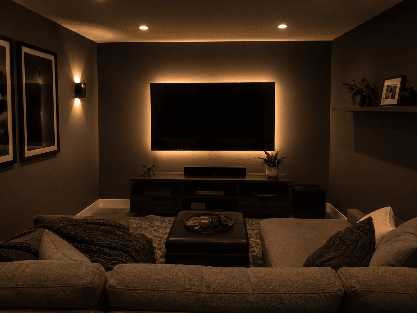 Small media room with a large wall-mounted TV, accent lighting behind the screen, sectional sofa and warm moody lighting.