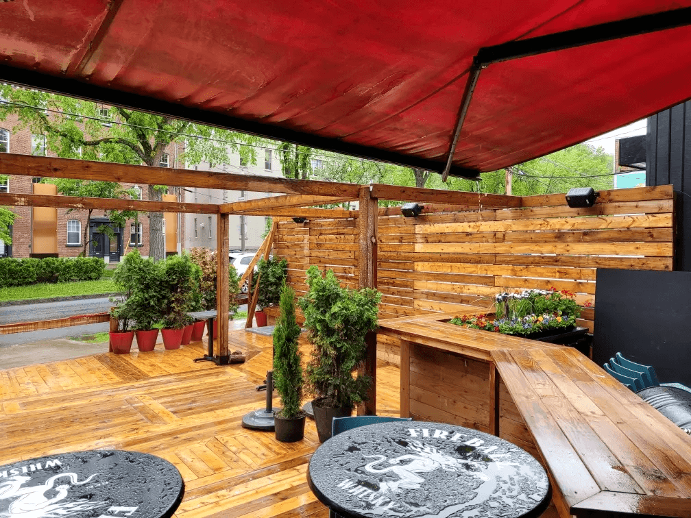 Commercial outdoor wooden terrace with pergola, red awning, horizontal slat privacy fencing, built-in bar counter, wooden decking, round tables, and decorative planters surrounded by greenery.
