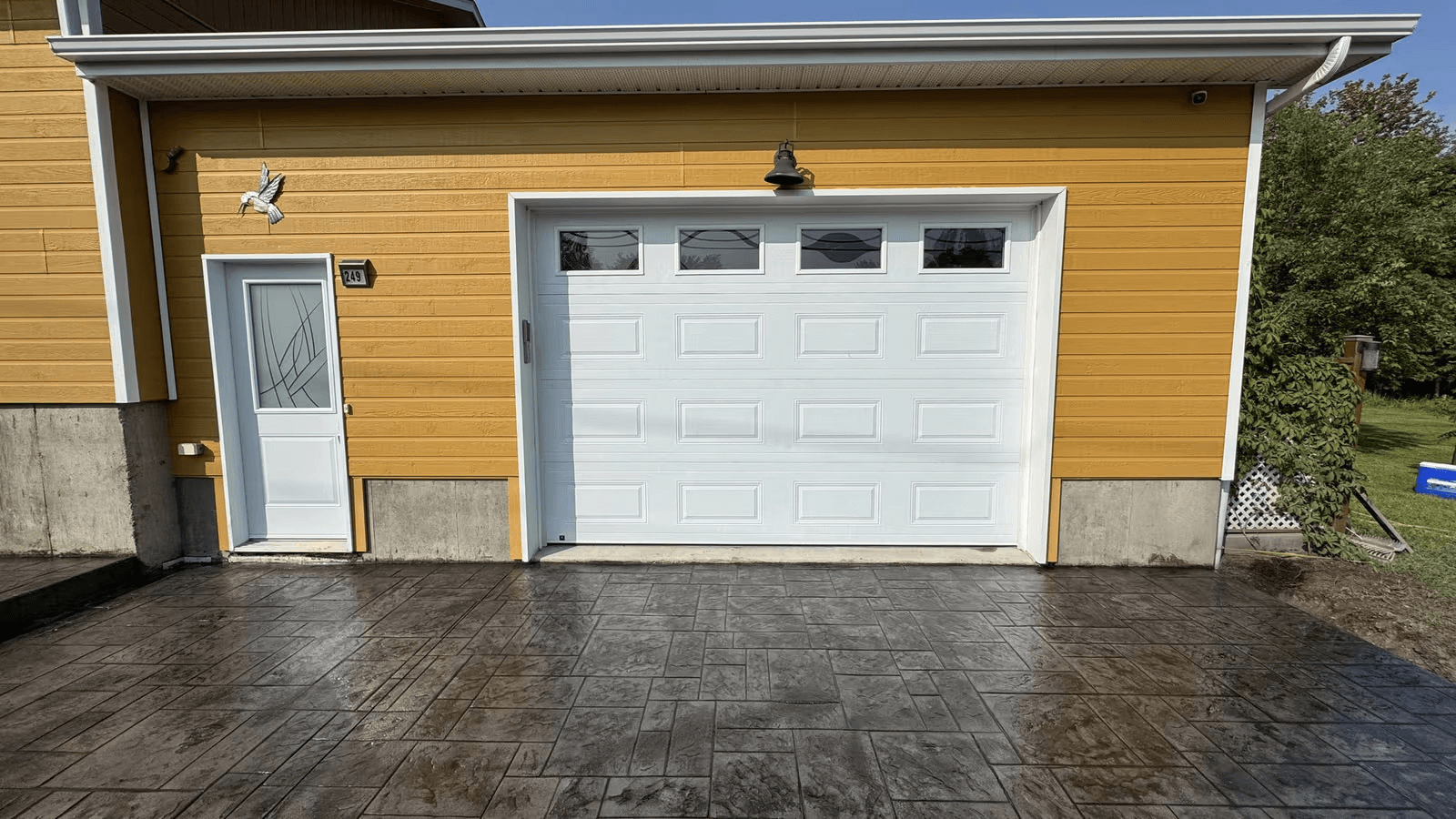 Residential garage facade with white garage door, yellow exterior siding, side entry door, soffits, fascias and stamped decorative concrete driveway.