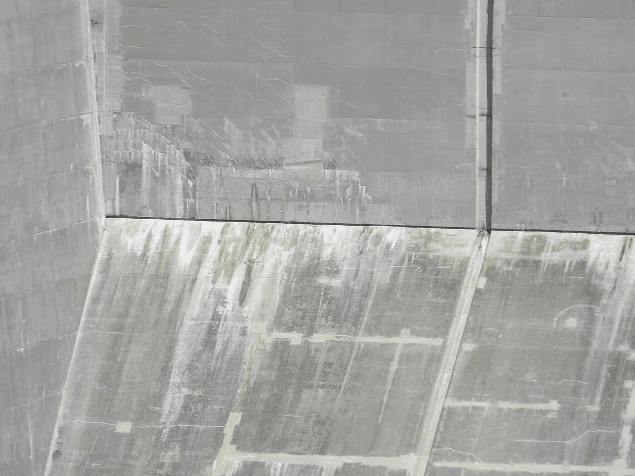 Concrete foundation wall showing fine cracks, moisture stains, streaking and mineral deposits on an inclined concrete surface.