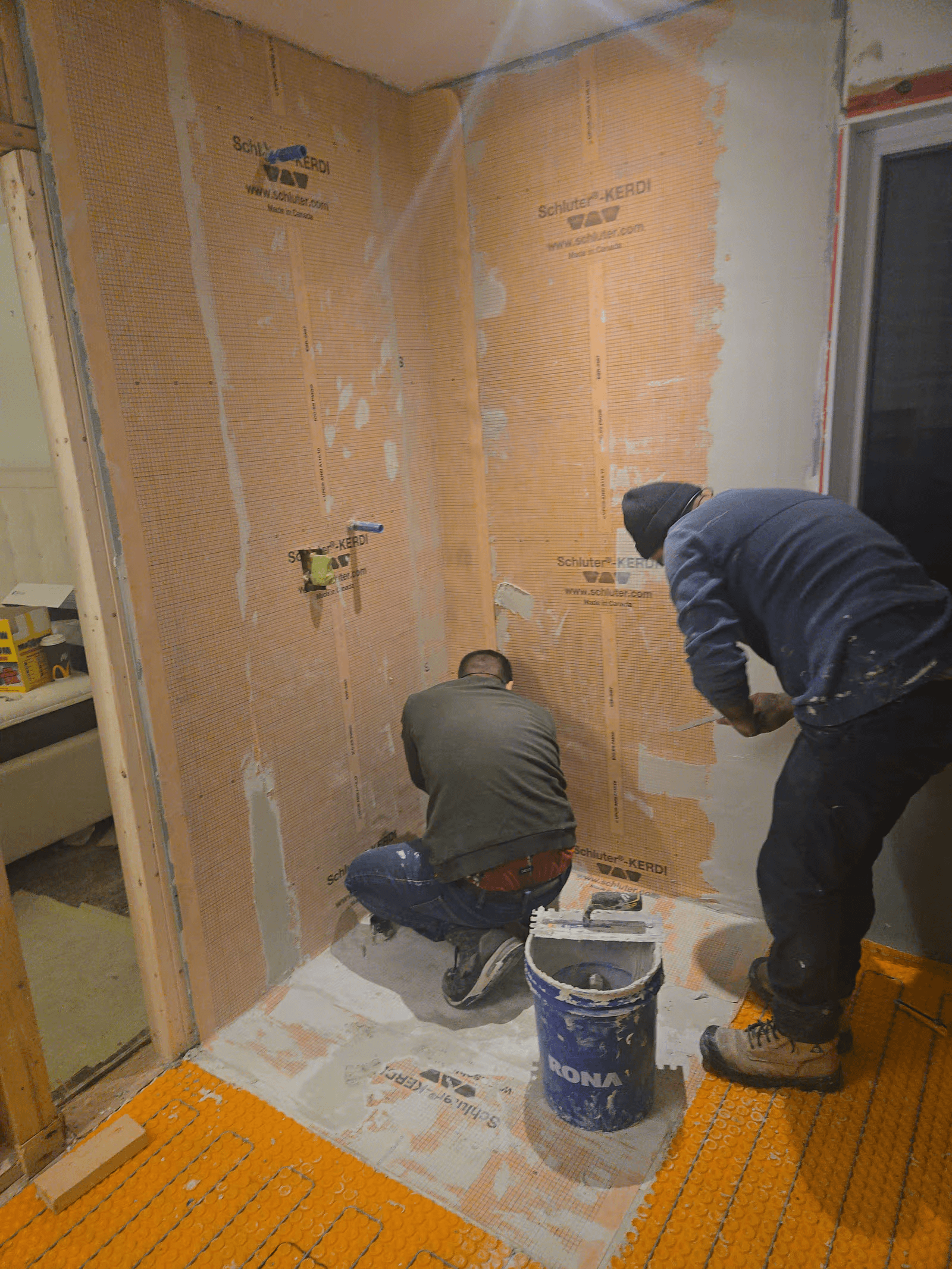 Two construction workers installing an orange waterproofing membrane in a shower during a renovation, preparing the walls and floor before tiling.