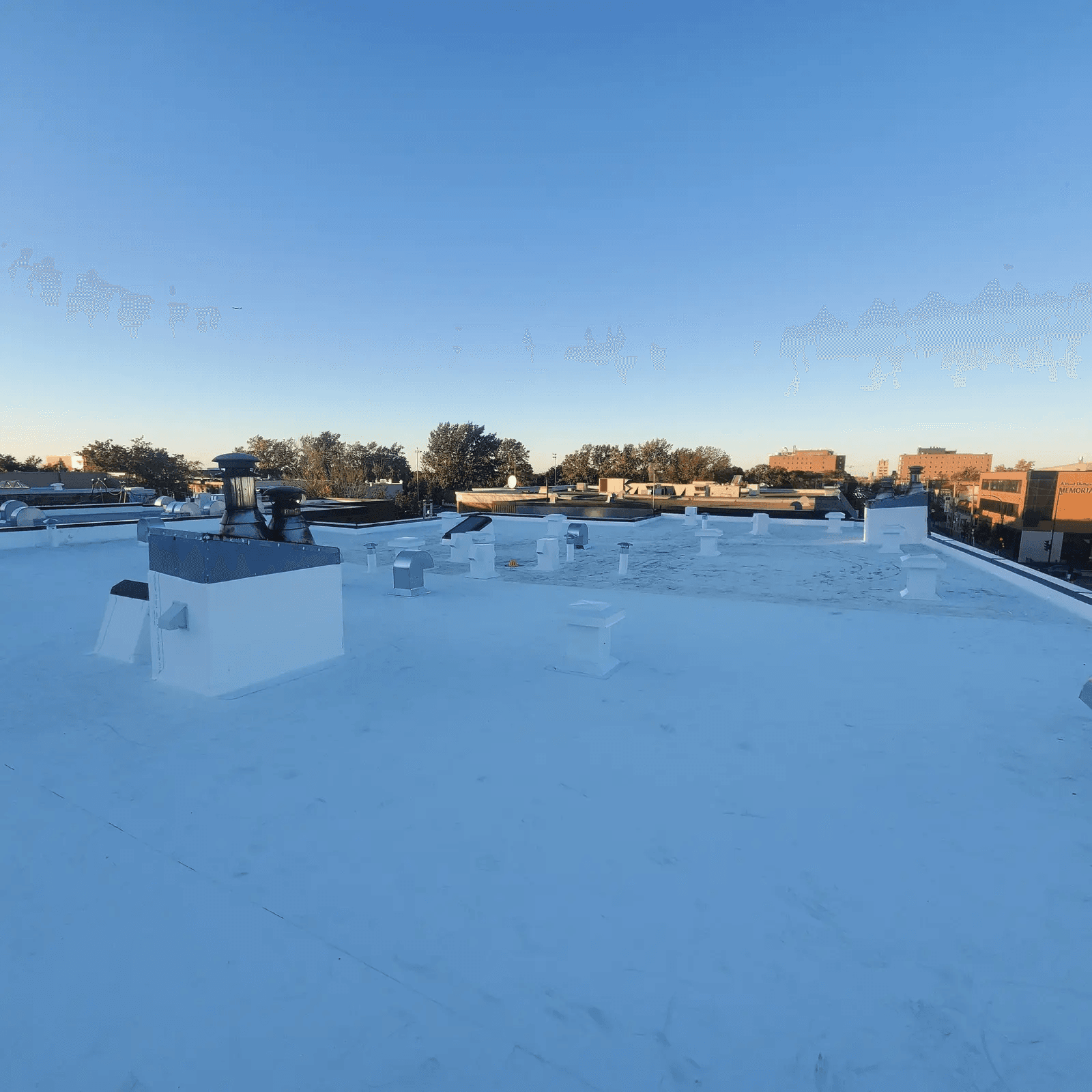 Large flat roof covered with a white reflective membrane featuring multiple vents, ventilation outlets and rooftop equipment in an urban setting