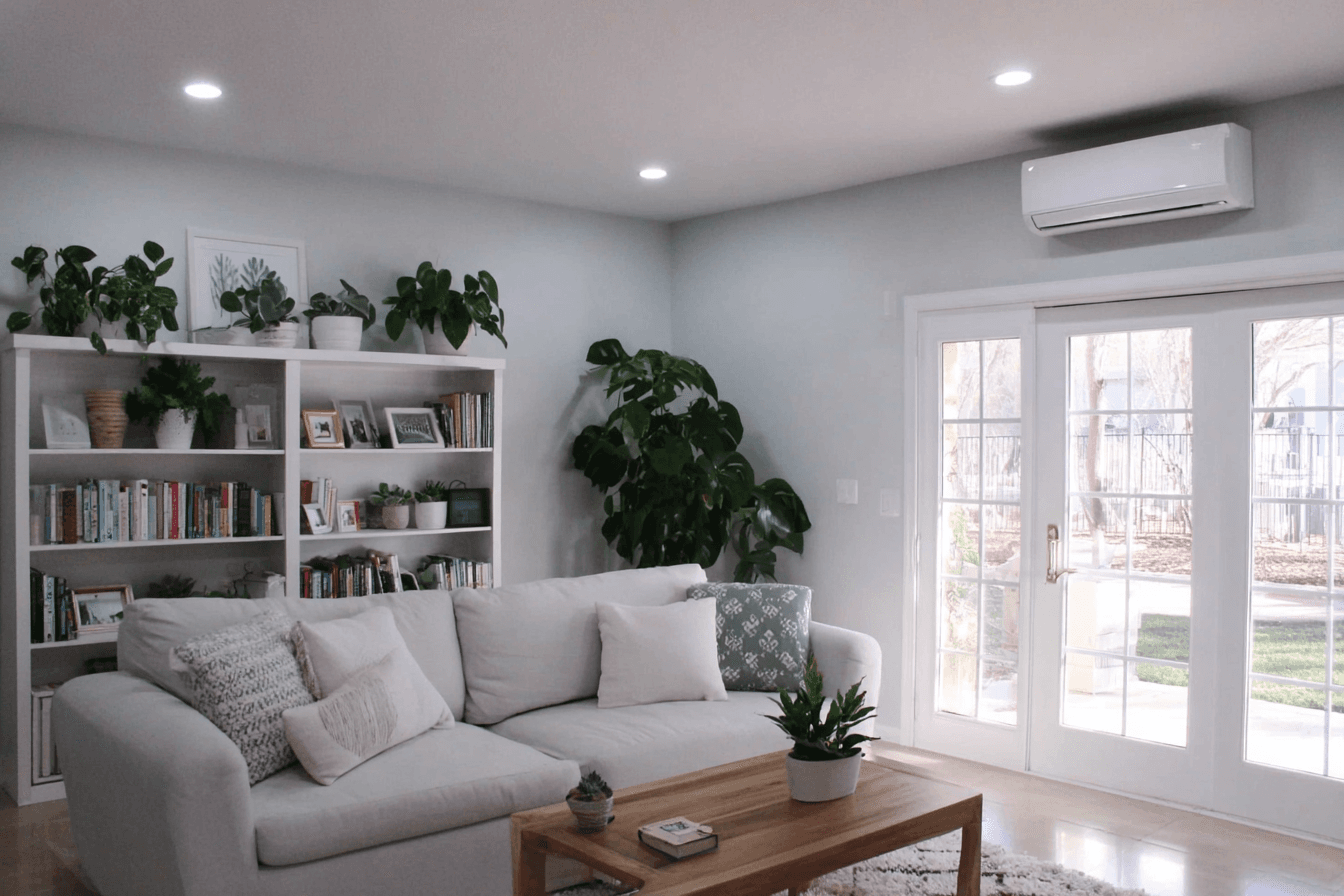 Bright living room with a white sectional sofa, plant-filled bookshelf, glass doors and a wall-mounted heat pump or air conditioning unit