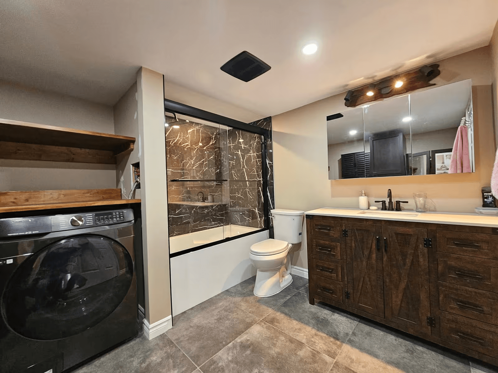 Renovated bathroom featuring a glass-enclosed shower with black marble-look tiles, dark wood vanity, double sink, grey tiled flooring and integrated laundry area.