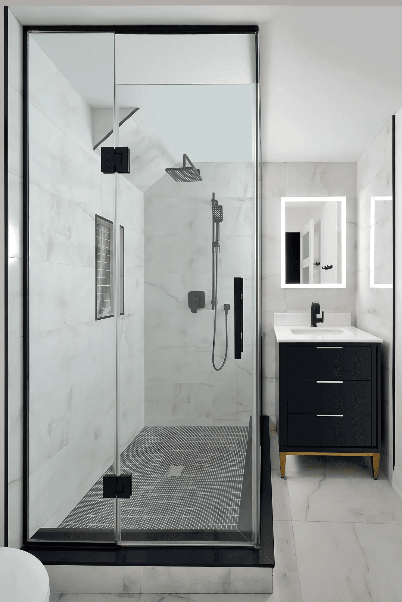 Modern renovated bathroom featuring a glass shower, white marble-look tiles, black fixtures, black vanity and illuminated mirror.