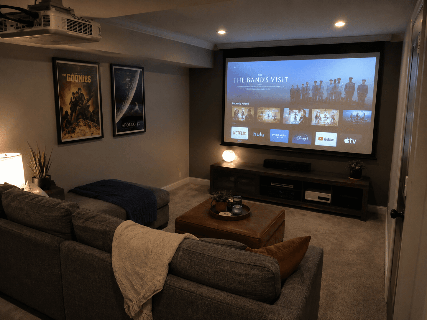 Home theatre with a ceiling-mounted projector, large projection screen, sectional sofa, media console and framed movie posters.