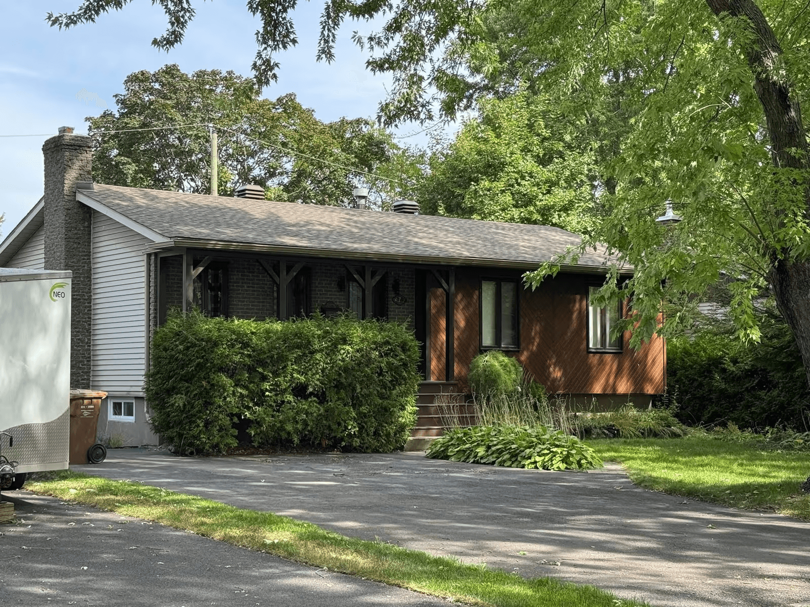 Single-storey detached house with a grey asphalt shingle roof, masonry chimney, brown wood and grey vinyl exterior cladding, covered porch with wood columns, rectangular windows, asphalt driveway and mature landscaping.