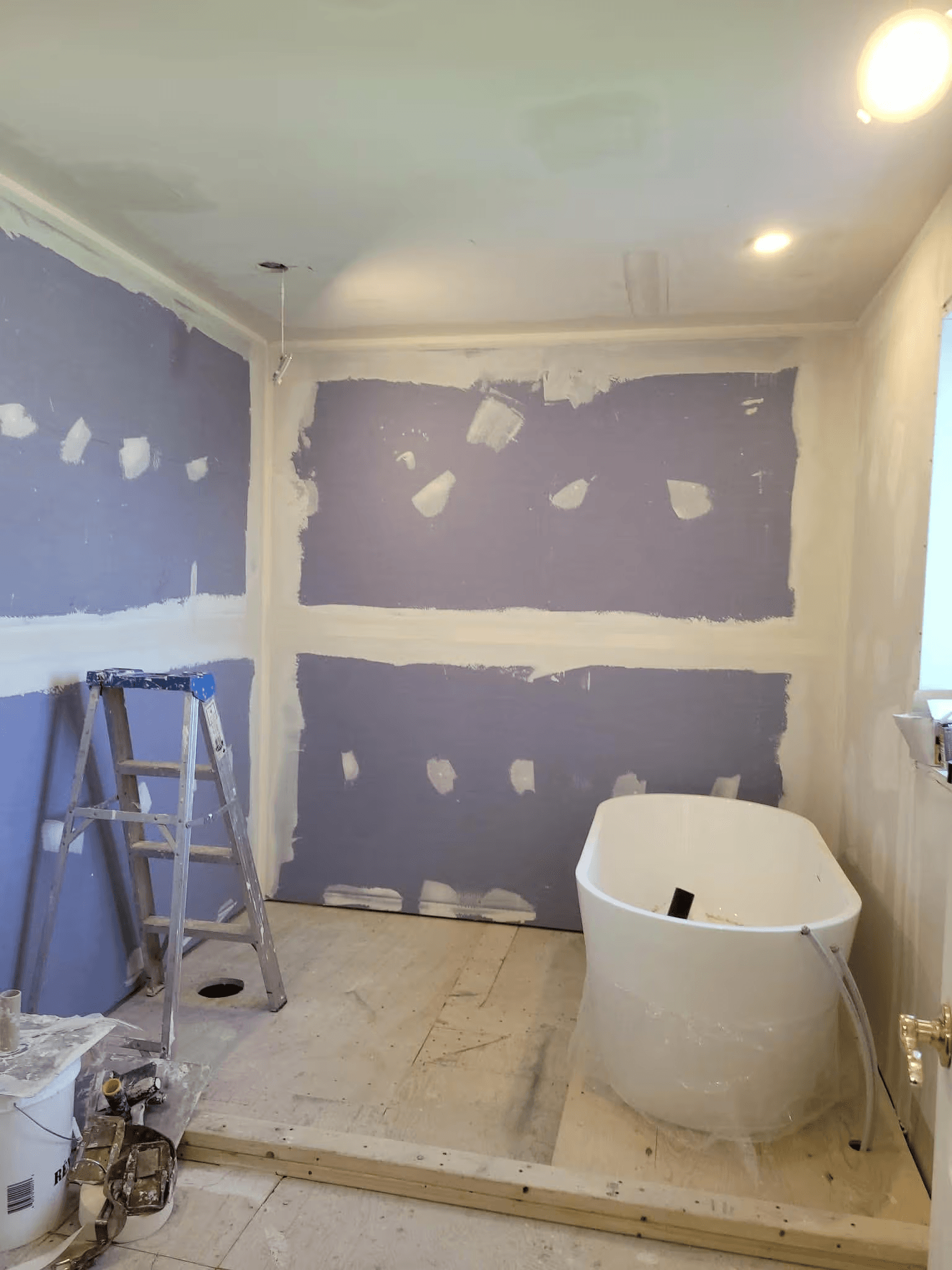 Bathroom renovation in progress featuring the installation of a white freestanding bathtub, purple drywall panels, joint compound application, ceiling repairs, plywood subfloor and construction stepladder