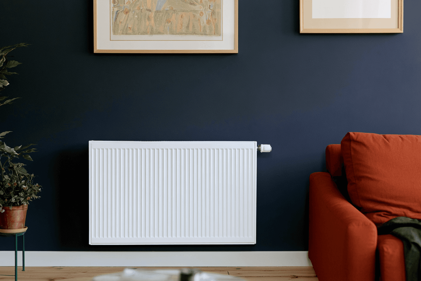 White wall-mounted radiator installed on a dark blue wall in a modern living room with an orange sofa, plants and hardwood flooring