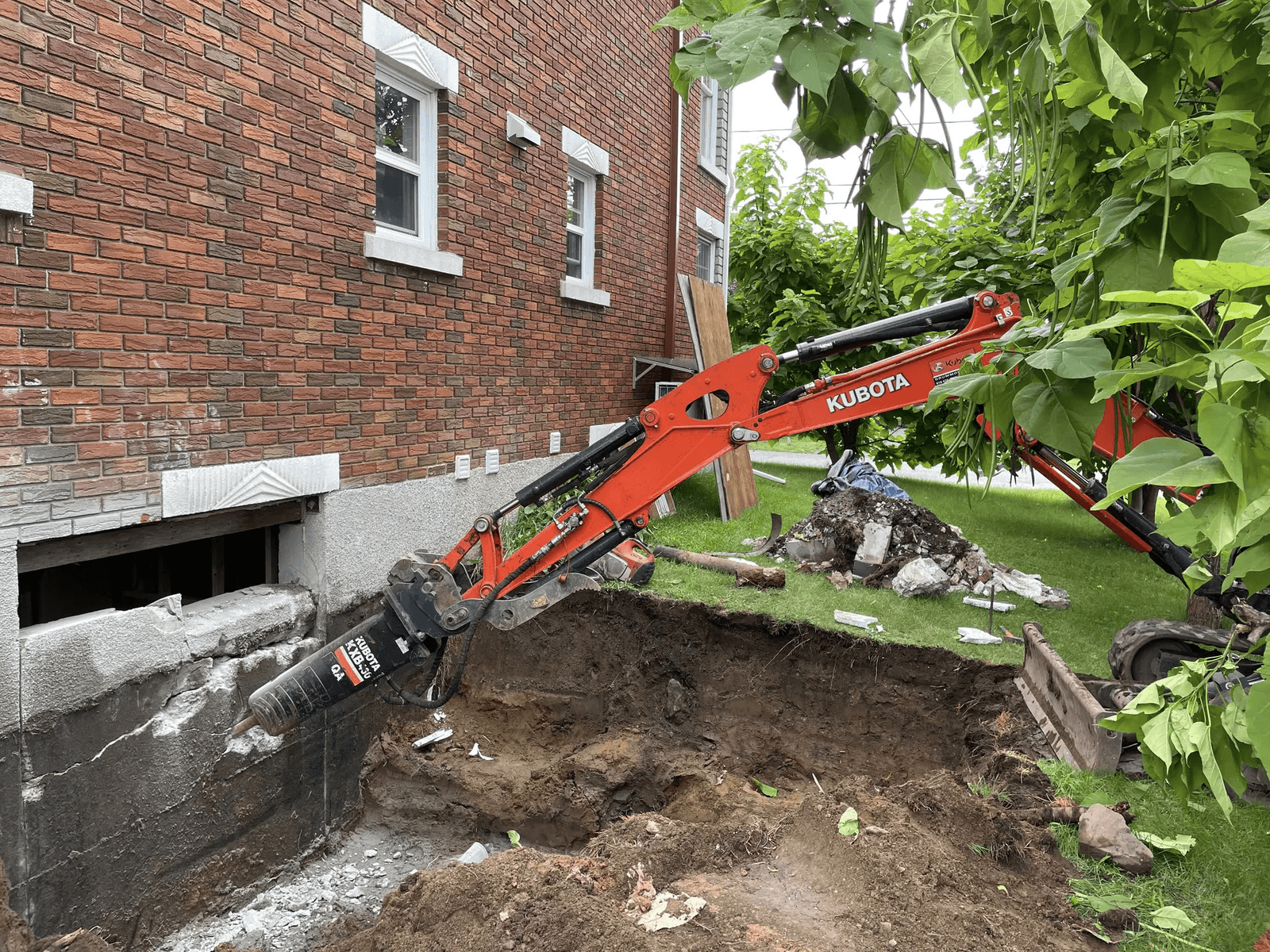 Kubota compact excavator equipped with a hydraulic breaker performing excavation and demolition work beside the foundation of a brick house.