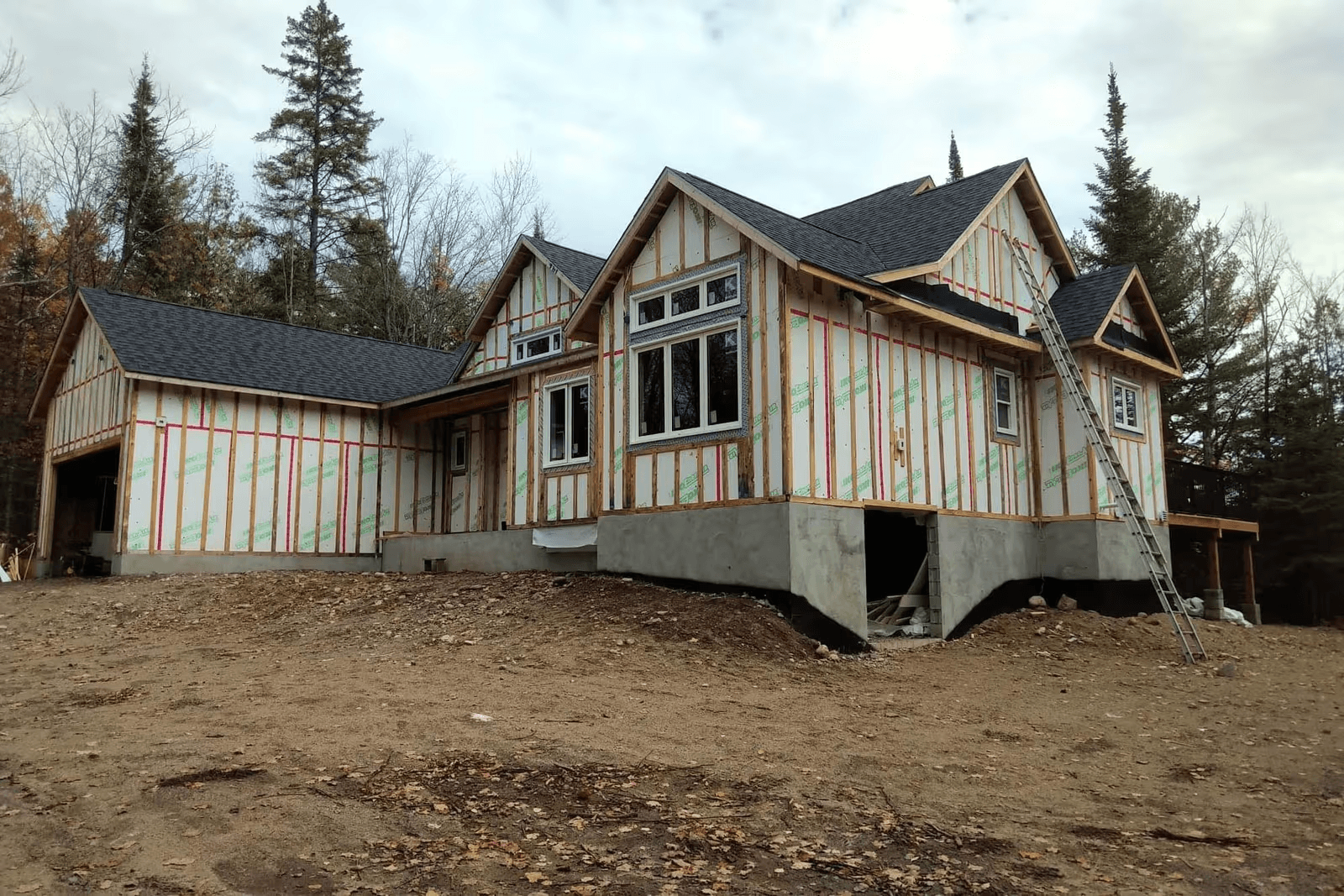 New detached house under construction with an attached garage on a wooded lot, featuring wood framing, insulated wall panels, black asphalt shingles, large windows and concrete foundations.