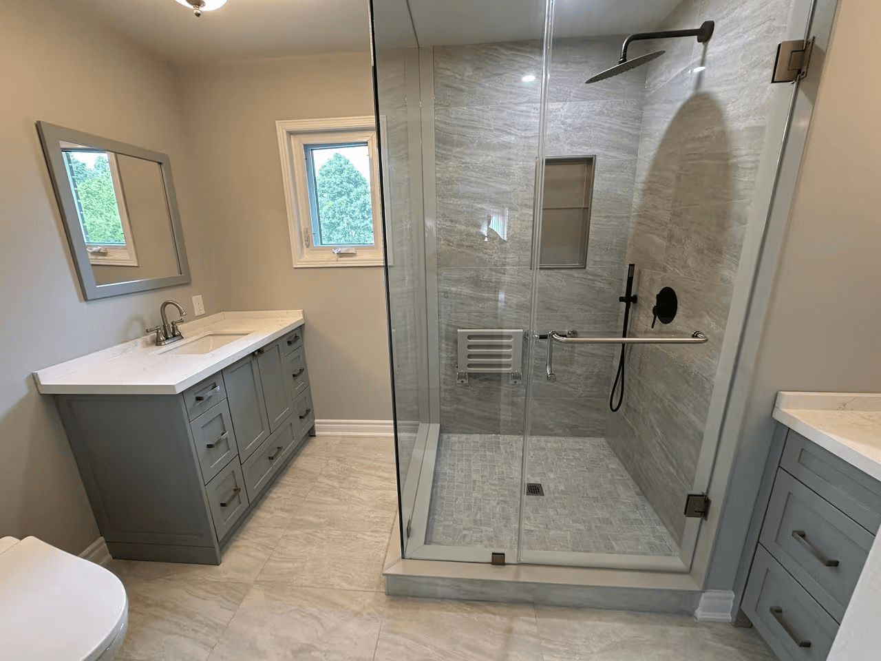 Renovated bathroom with a large glass-enclosed shower, grey ceramic tiles, black rainfall showerhead, grey vanities and white countertops.