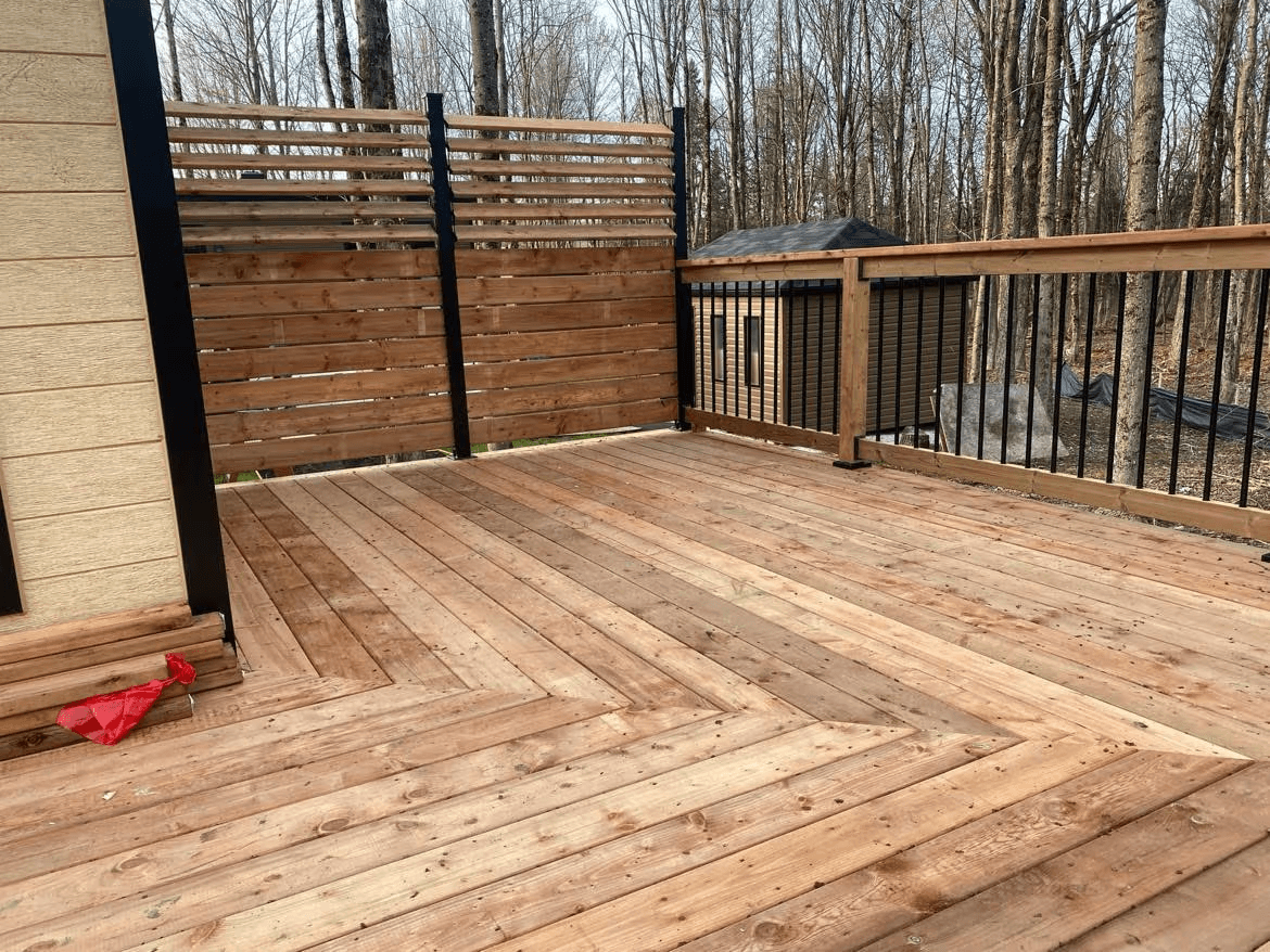 Large residential deck made of natural wood boards, featuring black metal railings and horizontal wooden privacy screens, located beside a beige-sided house and surrounded by mature trees.