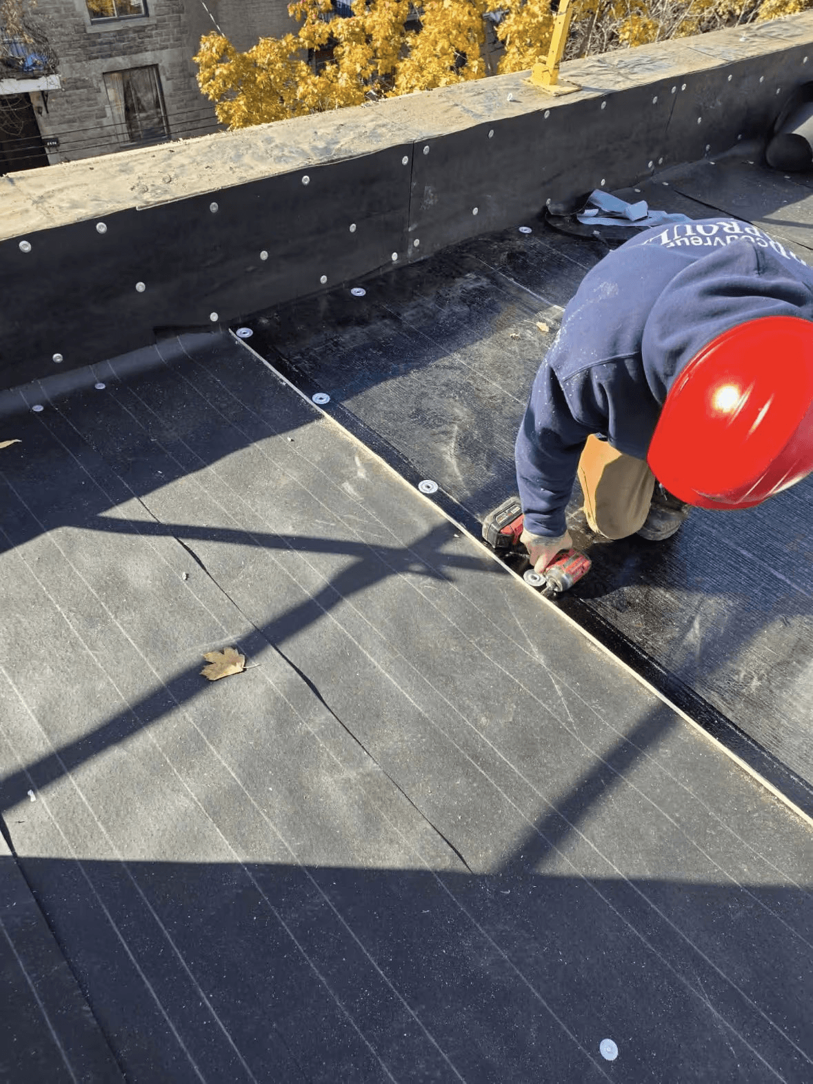 Worker installing and fastening a waterproofing membrane on a flat roof using a power tool.