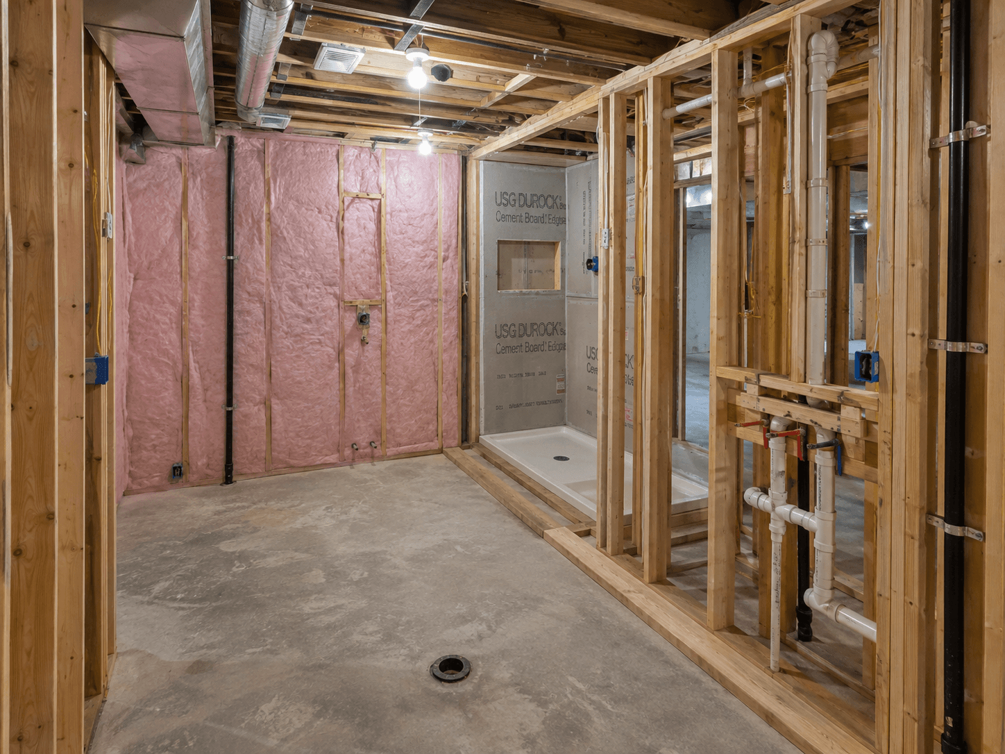 Basement bathroom under construction with wood framing, fiberglass insulation, exposed plumbing, ventilation ducts, and an installed shower base