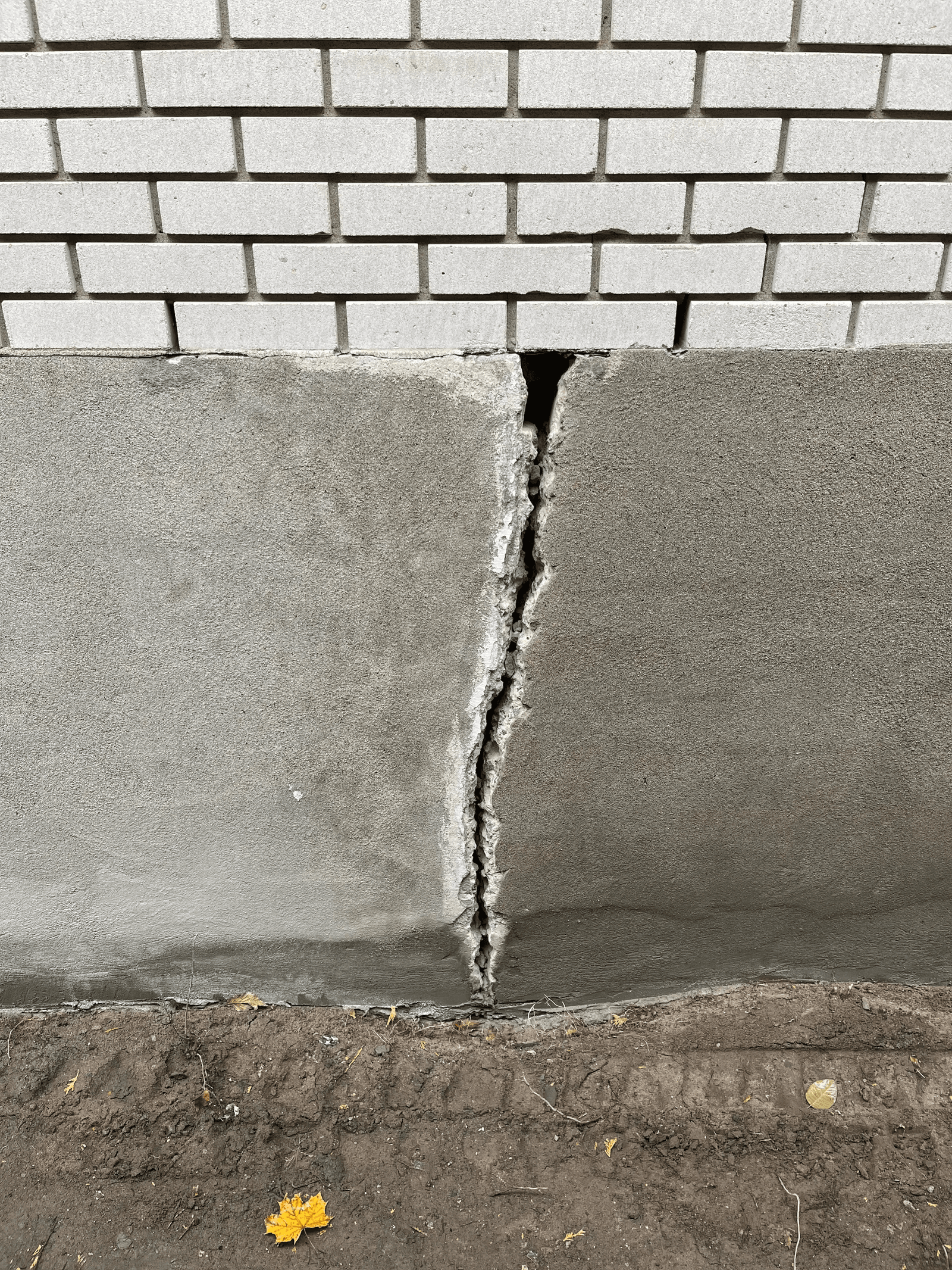 Large vertical crack in an exterior foundation wall beneath brick cladding, with damaged concrete requiring structural repair and waterproofing