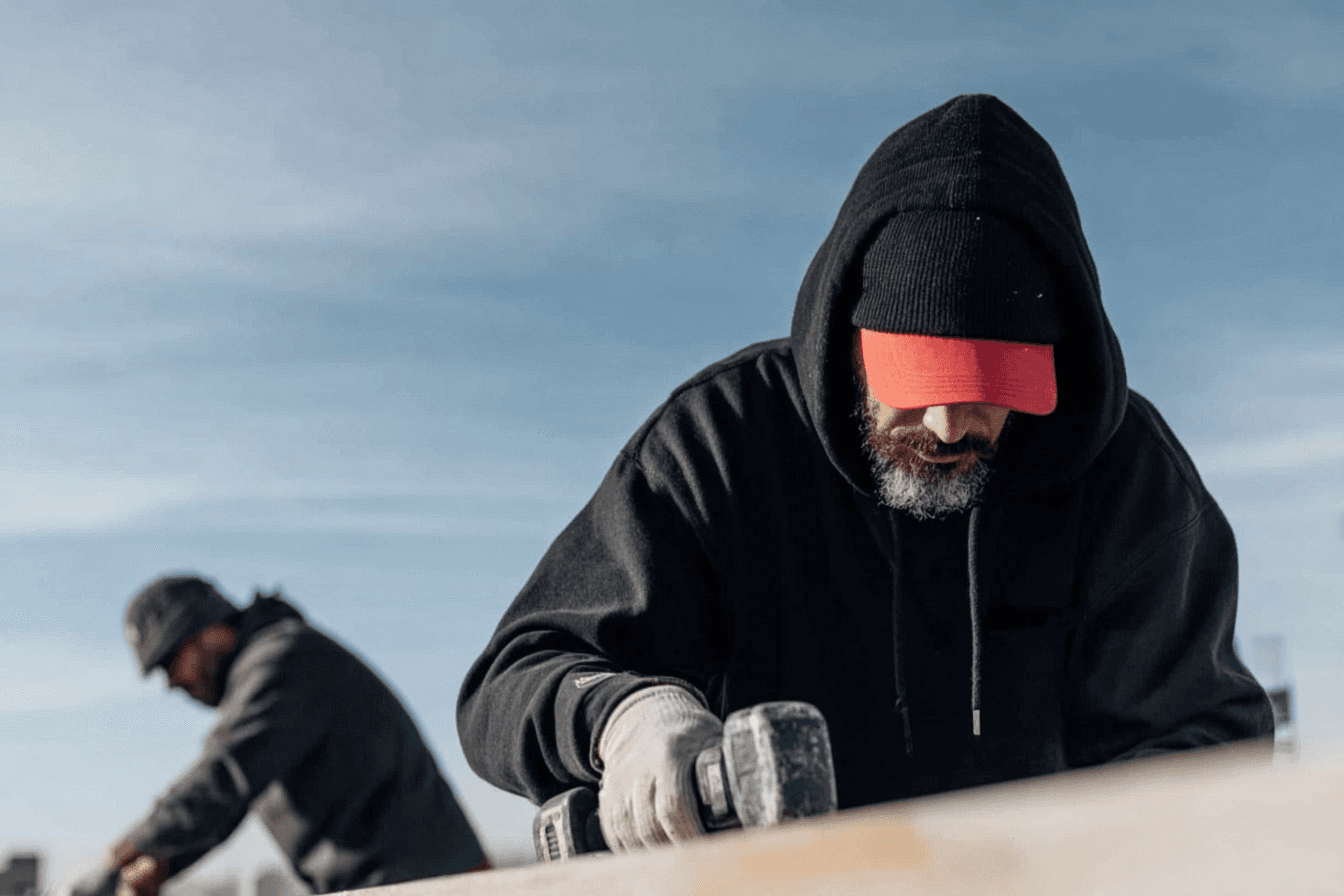 Construction worker using a power tool on a piece of wood, with a second worker in the background at an outdoor jobsite.