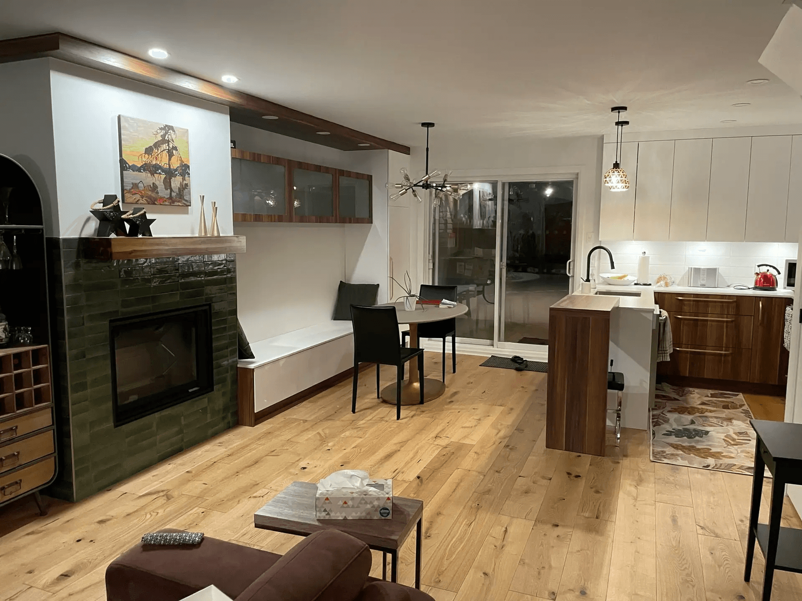 Renovated open-concept living space with a modern kitchen, built-in fireplace, wood flooring, dining area, and a large glass door opening to the outdoors.