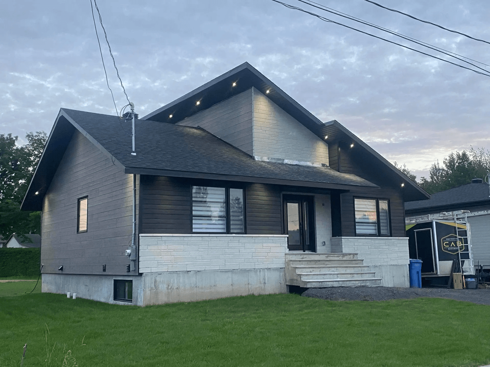 Modern house with dark wood siding and white stone on the lower part of the façade. It has a gable roof with recessed lighting beneath the eaves, concrete steps leading to the main entrance and a garage visible on the right.