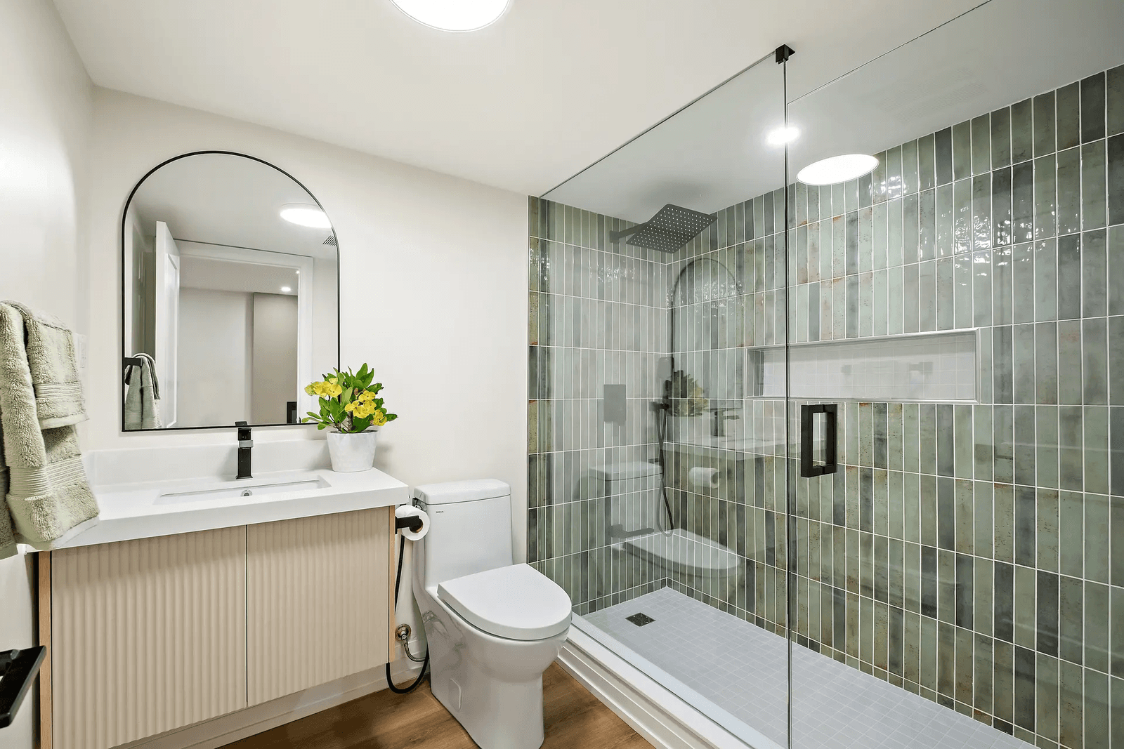 Modern bathroom featuring a glass shower, green wall tiles, light wood vanity, rounded mirror and elegant accessories.