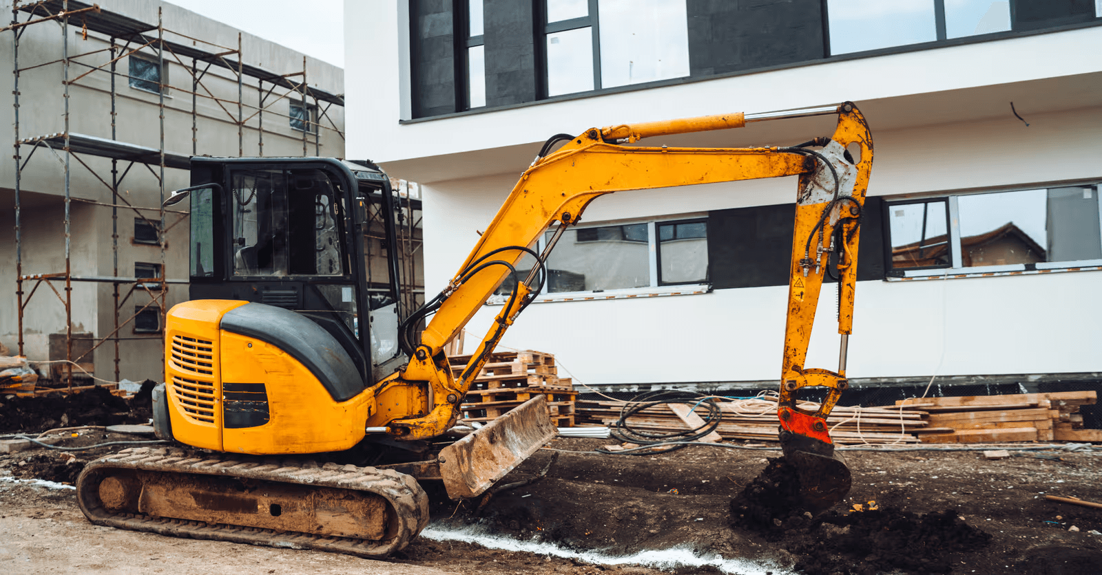 Compact tracked excavator performing earthmoving work in front of a modern residential building under construction, with scaffolding and building materials.