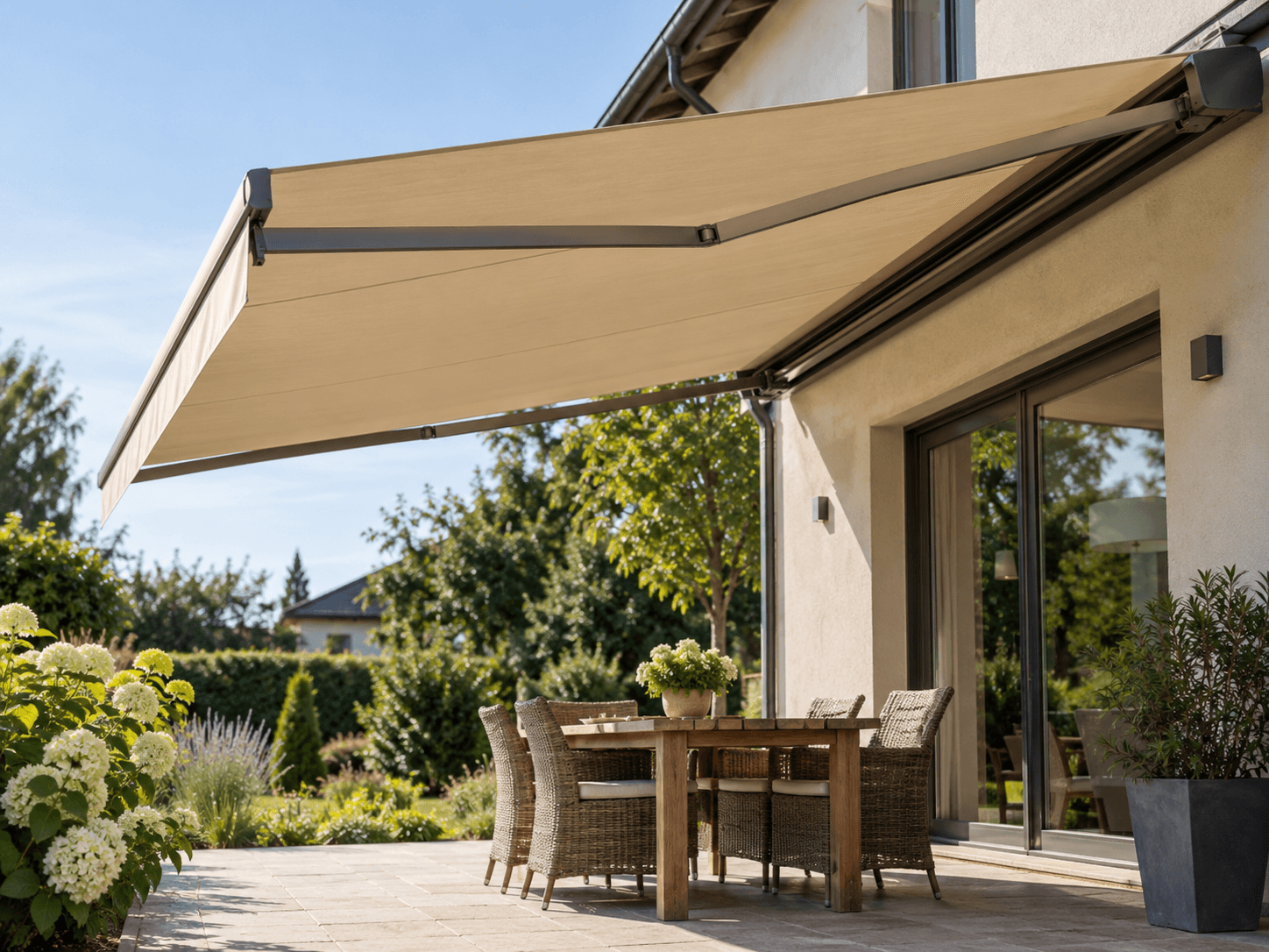 Modern retractable awning with new beige fabric and grey metal articulated arms, installed on a residential stucco exterior to create a shaded area above a tiled patio with outdoor furniture and a landscaped garden.