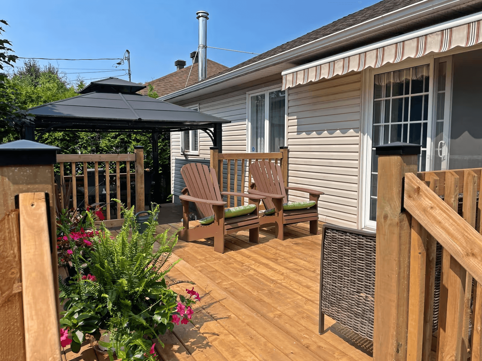 Natural wood residential deck with railings, two brown Adirondack chairs, grey gazebo, striped retractable awning, flower planters, and beige house siding with a glass patio door.
