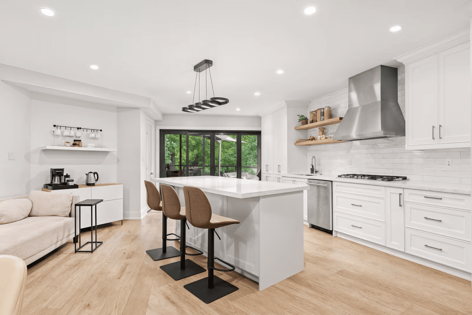 White, modern kitchen with a wide-span island, stools, stainless steel range hood, custom-built cabinets and light wooden floors.