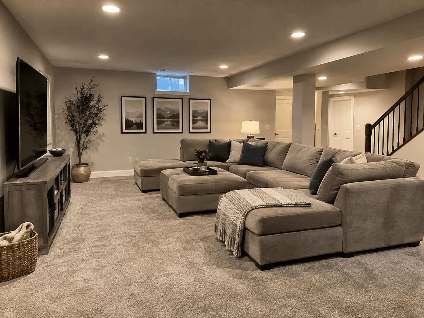 Spacious finished basement with gray sectional sofa, beige carpet, wooden TV console, recessed lighting, and open staircase.