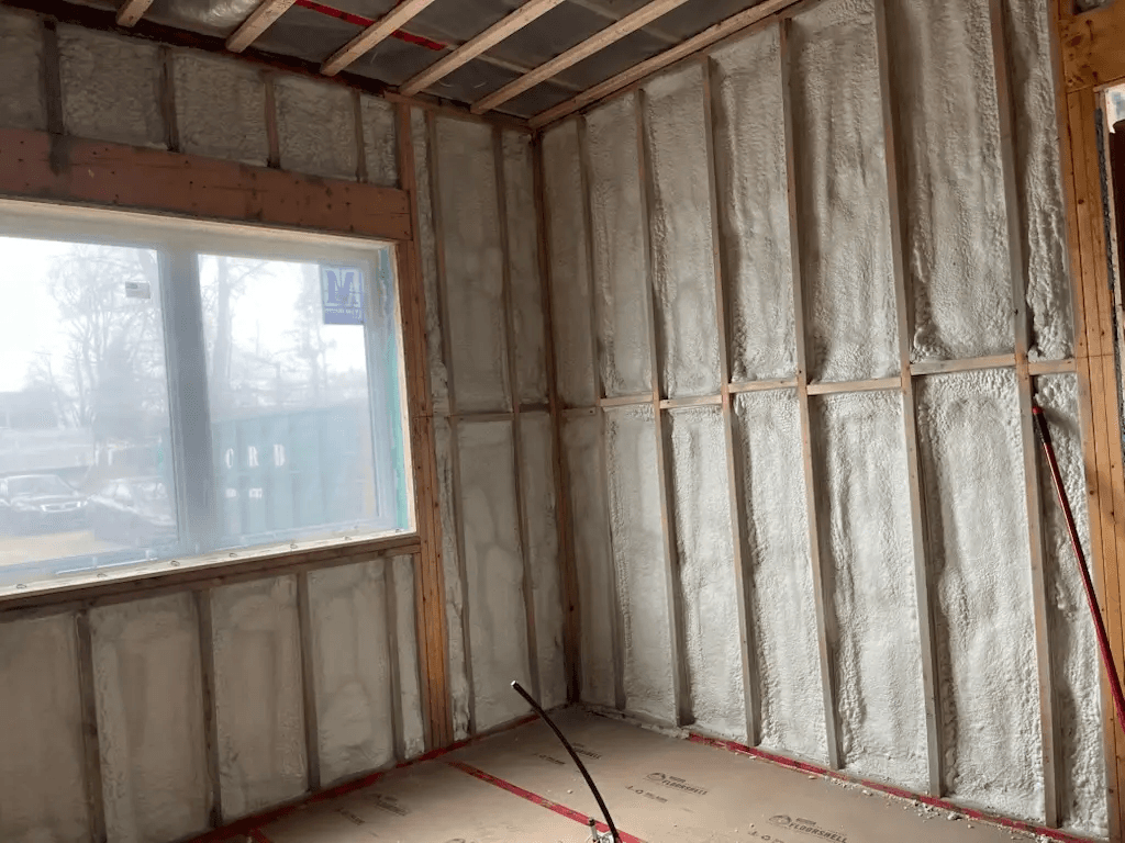 Interior wall under renovation with wood framing, spray foam insulation, an installed window, and ceiling preparation before drywall installation