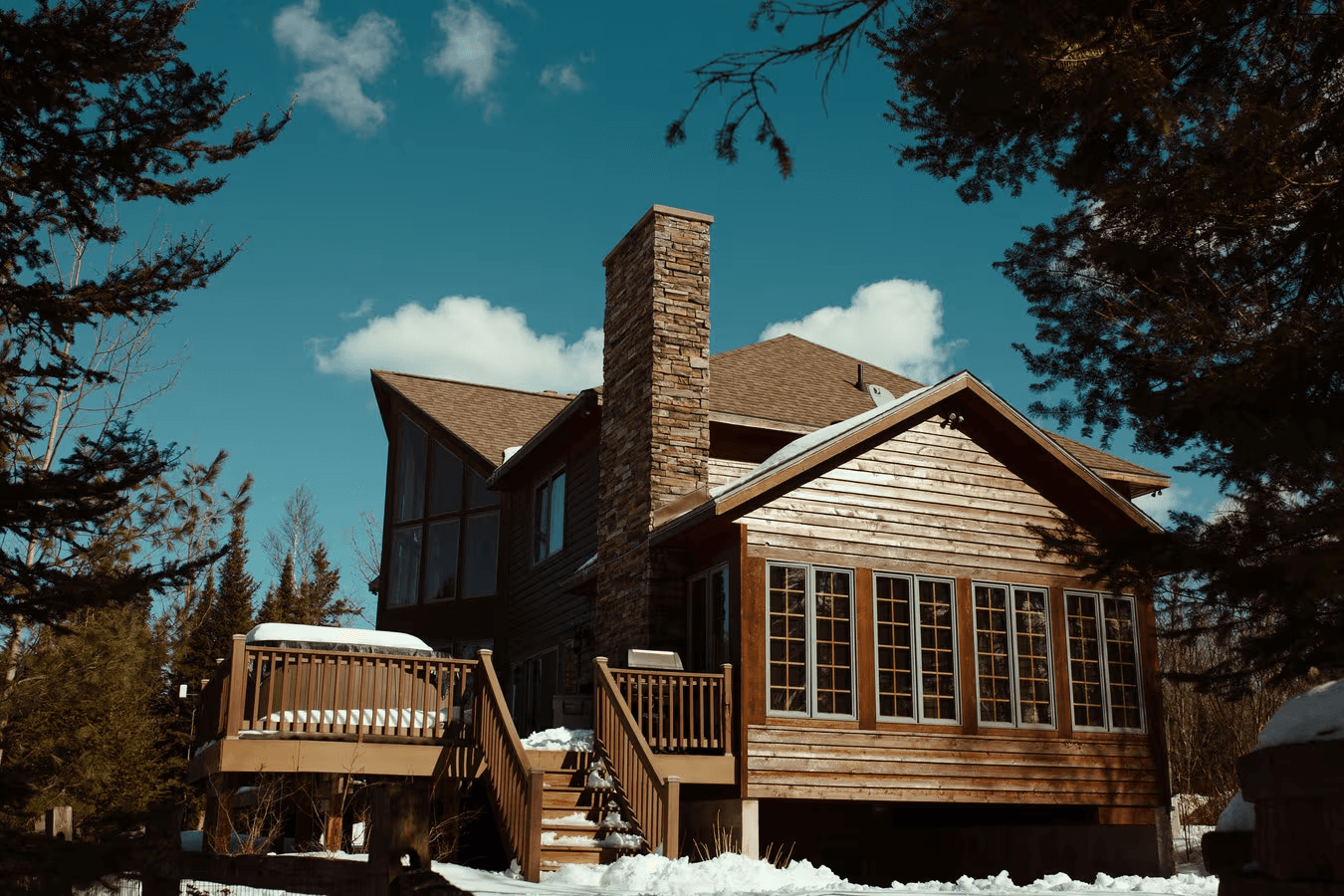 Wood-sided residential house with a shingle roof, large stone chimney, rear deck, exterior stairs and multiple windows in a snowy wooded setting.