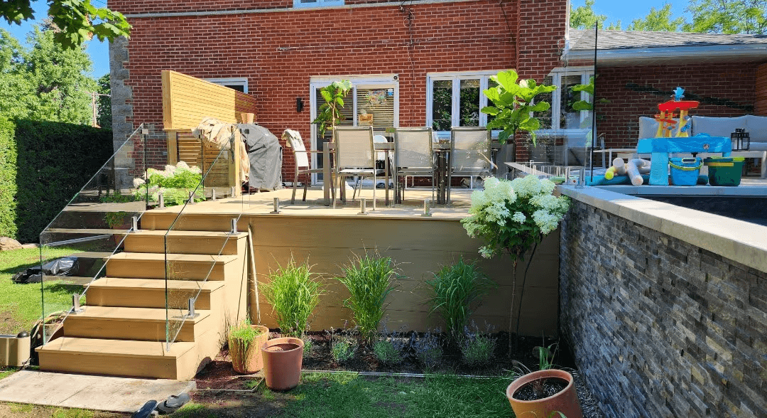 Elevated residential wooden deck with wide stairs, transparent glass railings, patio furniture, wooden privacy screens and landscaping with ornamental grasses and hydrangeas, located beside a swimming pool, a low grey stone wall and a red brick house.