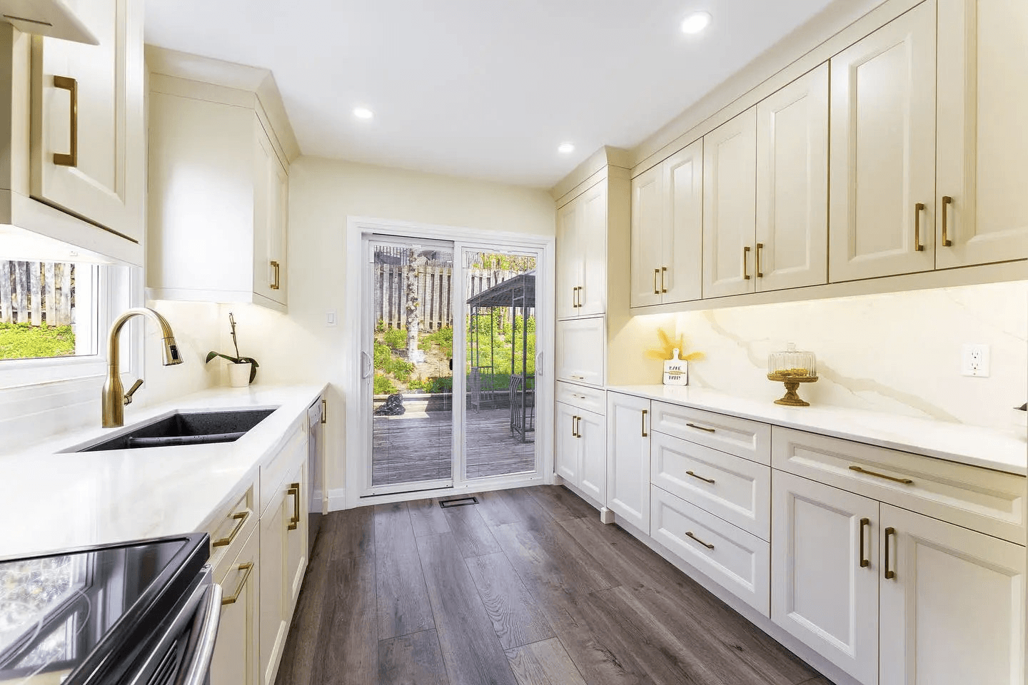 Bright modern kitchen with white cabinets, light countertops, black sink, metal faucet, wood flooring and a glass patio door opening onto a deck.