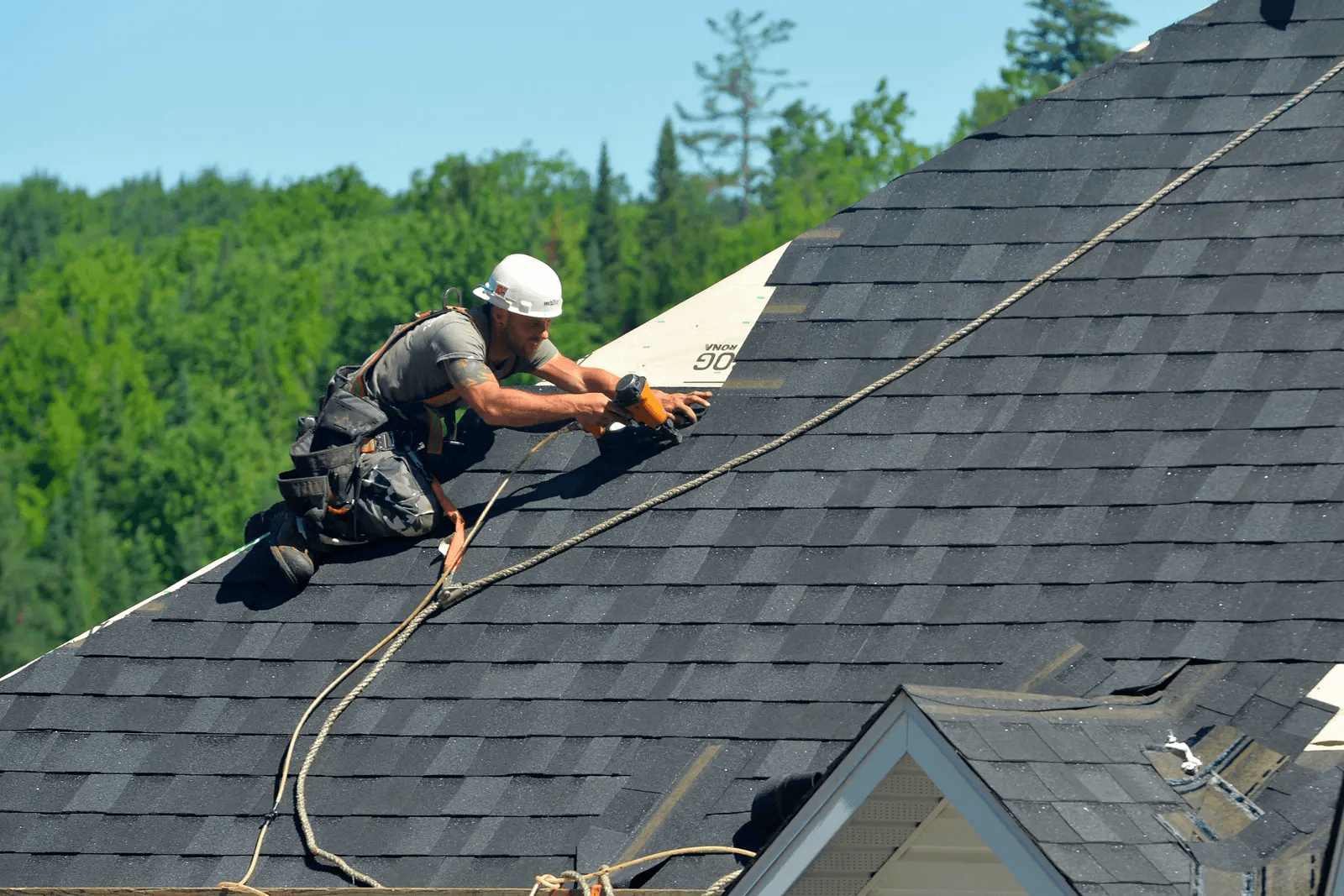 Worker on a sloped roof installing asphalt shingles with a roofing nail gun outdoors