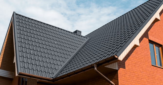 Residential dark charcoal gray metal tile roof with wavy profile, roof ridges, valleys, gutters and downspouts on a modern red brick house with gables and roof overhangs.