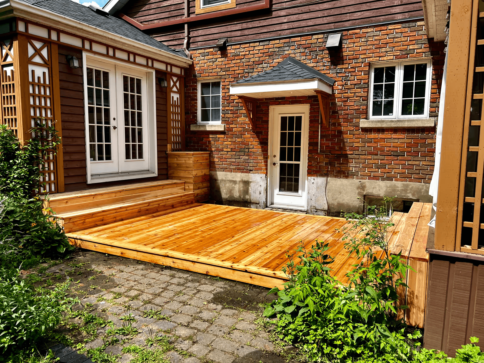 Residential outdoor deck made of natural wood, featuring new decking boards, wide steps, white French doors, decorative wooden lattice panels and landscaping in a courtyard surrounded by red brick walls and brown siding.