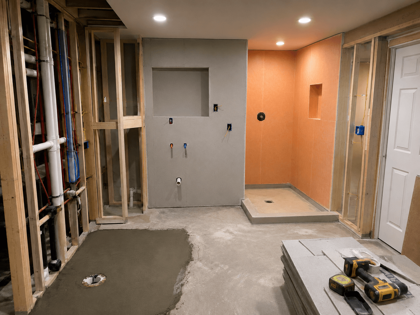 Basement bathroom renovation in progress with wood framing, exposed plumbing, recessed lighting, waterproof wall panels, and a shower base ready for tiling