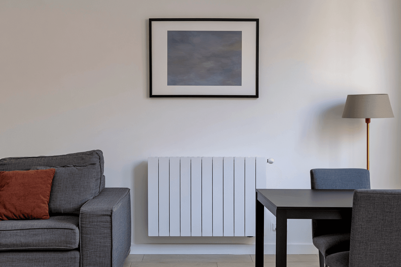 Modern white wall-mounted radiator installed in a living room with grey sofa, dining table, chairs and floor lamp