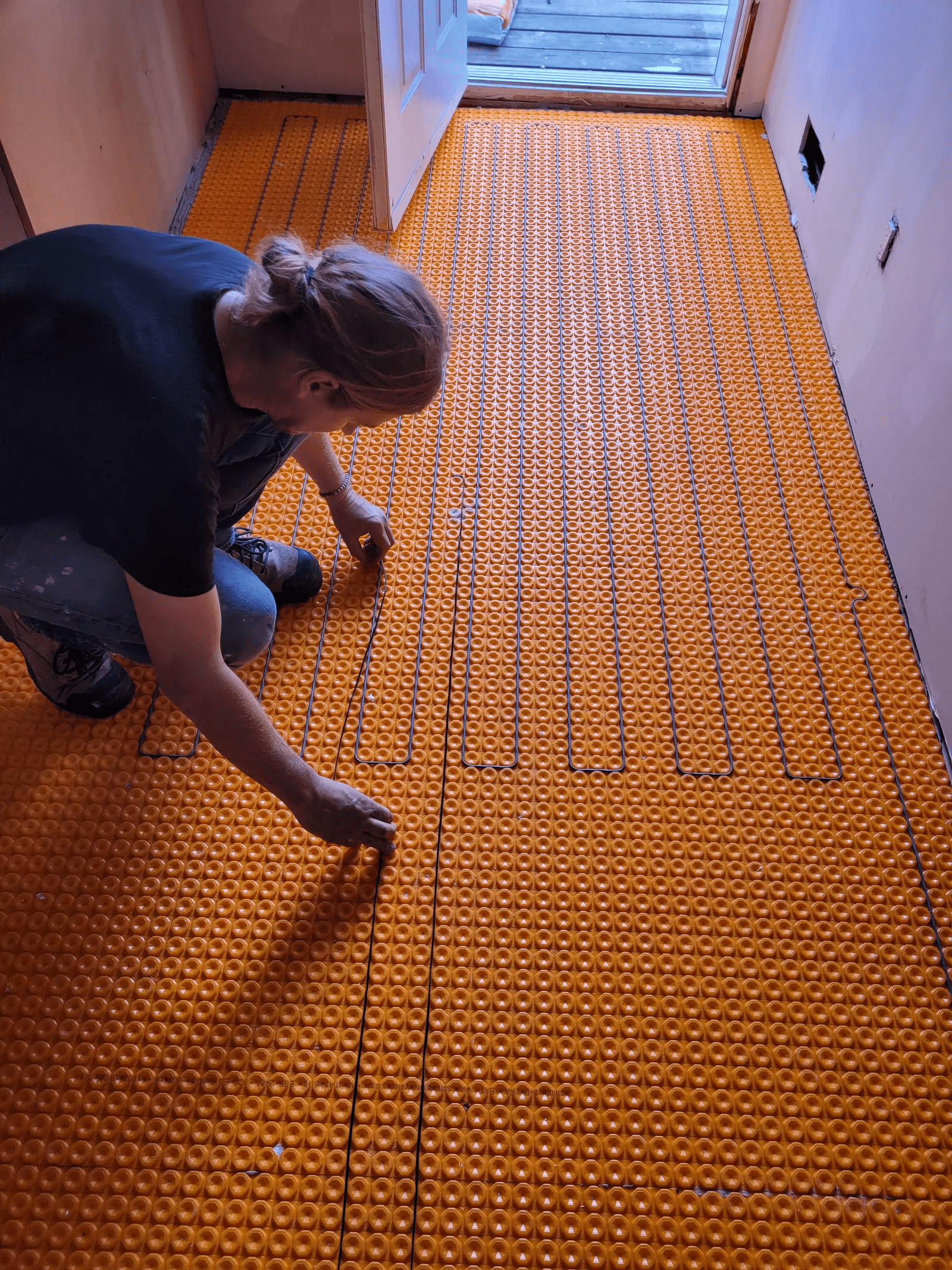 Worker installing an electric underfloor heating system with an orange membrane in a room under construction.